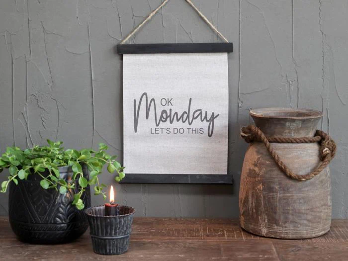 På væggen hænger et indrammet skilt med teksten »OK Monday LETS DO THIS« ved siden af en potteplante, et tændt stearinlys og en rustik kande med et rebhåndtag.