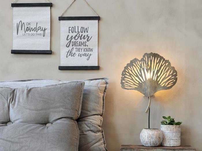 Hyggeligt opholdsrum med en tuftet grå sofa, to motiverende vægtæpper, en bladformet bordlampe og små potteplanter på et træbord.