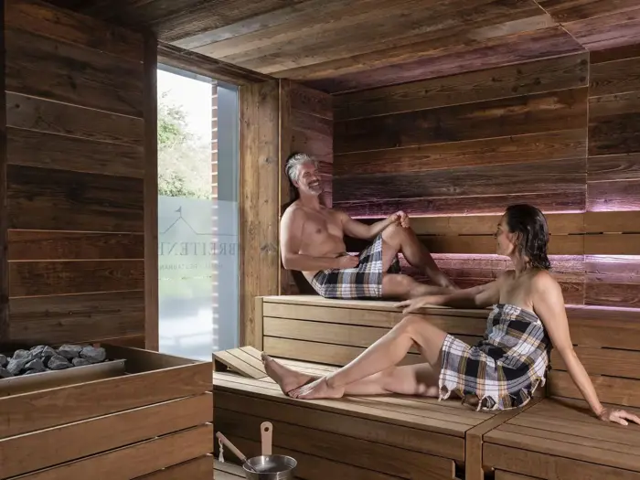 En mand og en kvinde i ternede håndklæder slapper af og smiler, mens de sidder på træbænke inde i en moderne sauna med naturligt lys, der kommer ind gennem et vindue.