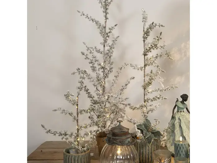 Tre små kunstige sneklædte træer i grønne potter er arrangeret på en træflade med stearinlys, en glaslygte og en englefigur.