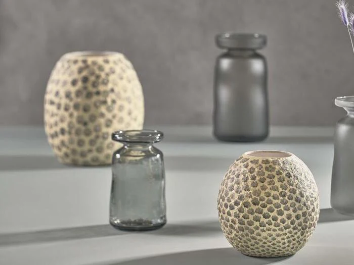 Fire dekorative vaser på en lys overflade; to er runde og strukturerede med et plettet mønster, to er cylindriske og lavet af gennemskinneligt røgfarvet glas.