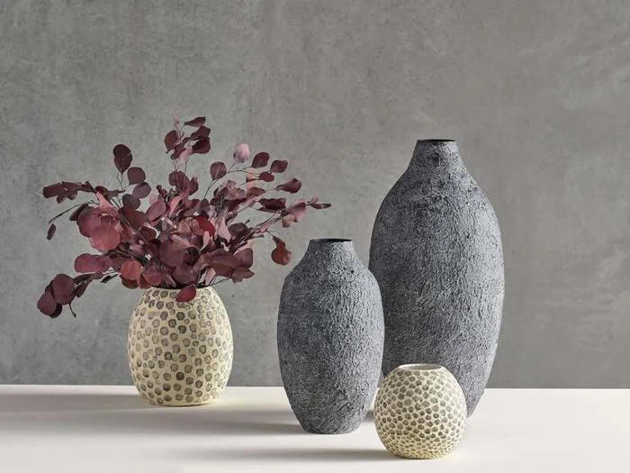 Tre strukturerede grå vaser og en cremefarvet vase med prikket mønster på et bord; den cremefarvede vase indeholder mørkerøde eukalyptusgrene.