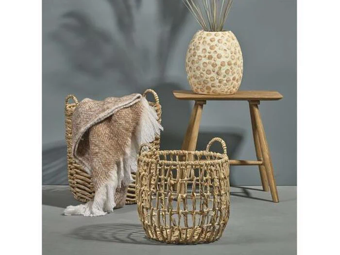 To flettede kurve, den ene med et brunt og hvidt tæppe, står ved siden af en træskammel med en dekorativ vase med høje stængler op ad en grå væg.