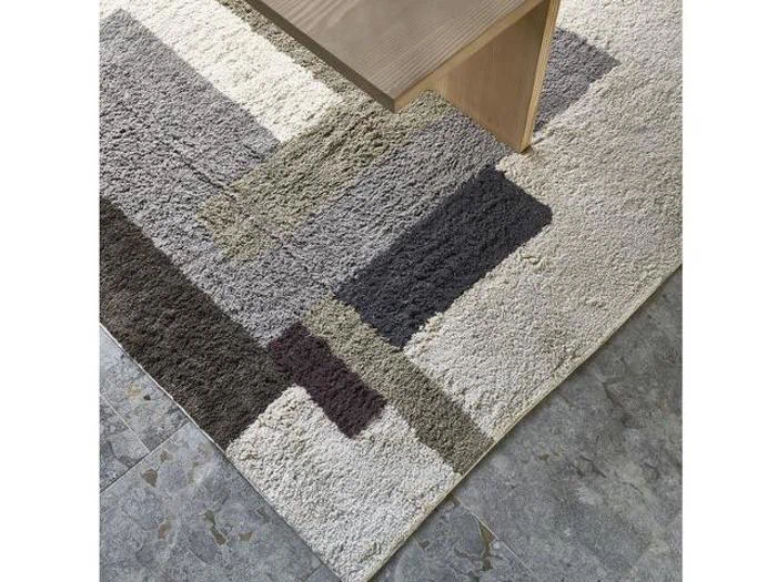 Et beige og gråt tæppe med rektangulære mønstre ligger delvist under et lyst træbord på et gråt klinkegulv.