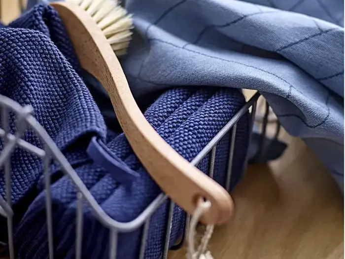 En opvaskebørste af træ og blå håndklæder er placeret i en metaltrådkurv på en træoverflade med et lyseblåt klæde i baggrunden.