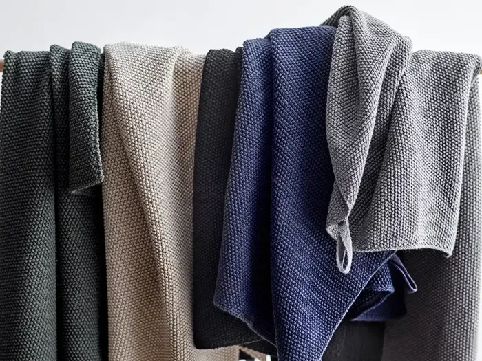 Fire strukturerede, strikkede tæpper i mørkegrå, beige, marineblå og lysegrå draperet over en sort skinne mod en almindelig, lys baggrund.