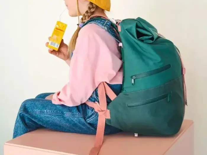 En person i lyserød sweater, blå overalls og gul hat sidder på en bænk med en grøn rygsæk på og drikker af en juicekarton med sugerør.