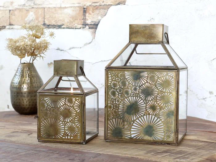 To dekorative guldlygter med indviklede geometriske mønstre står på et træbord. En messingvase med tørrede blomster står i baggrunden mod en hvid, let forvitret væg.