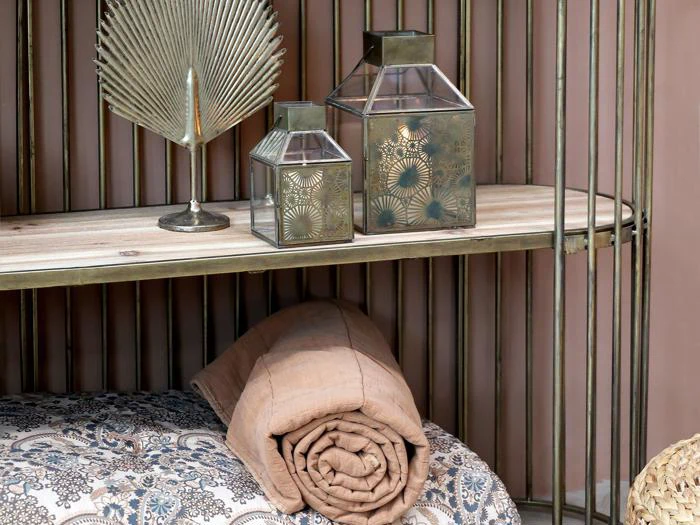 En hylde med to dekorative lanterner, et bladornament i metal, et sammenrullet beige tæppe og en mønstret pude nedenunder.