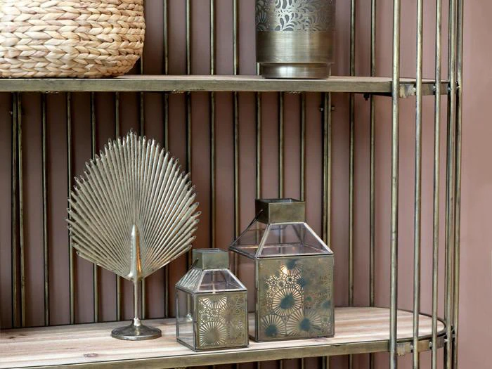 Metalhylde med en flettet kurv, en dekorativ vifteskulptur, to prægede lanterner og en cylindrisk vase mod en lyserød væg.