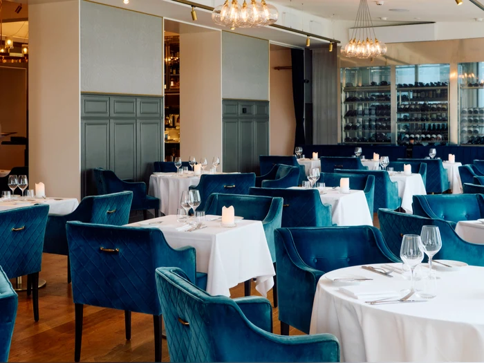 Elegant restaurantinteriør med blå fløjlsstole, hvide duge, dækkede borde, vinglas og blød belysning fra moderne lysekroner.