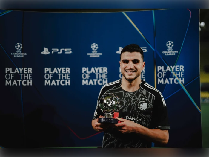 En smilende fodboldspiller i en sort trøje holder en pokal foran en baggrund med teksten PLAYER OF THE MATCH ved et UEFA Champions League-arrangement.