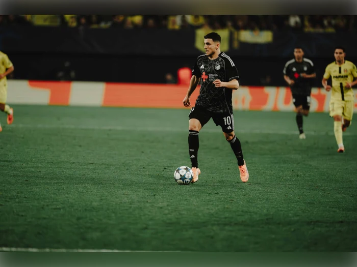 En fodboldspiller i sort uniform dribler bolden på en bane under en kamp, med andre spillere og et sløret publikum i baggrunden.