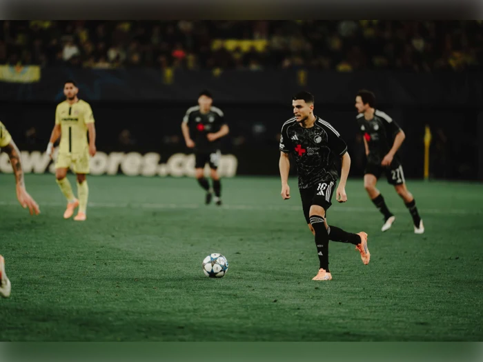 En fodboldspiller i sort uniform dribler med bolden under en kamp, mens holdkammerater og modstandere i gule uniformer ses i baggrunden.