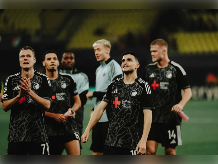 En gruppe mandlige fodboldspillere i sorte trøjer står sammen på en bane, nogle klapper og smiler efter en kamp. Baggrunden er sløret.