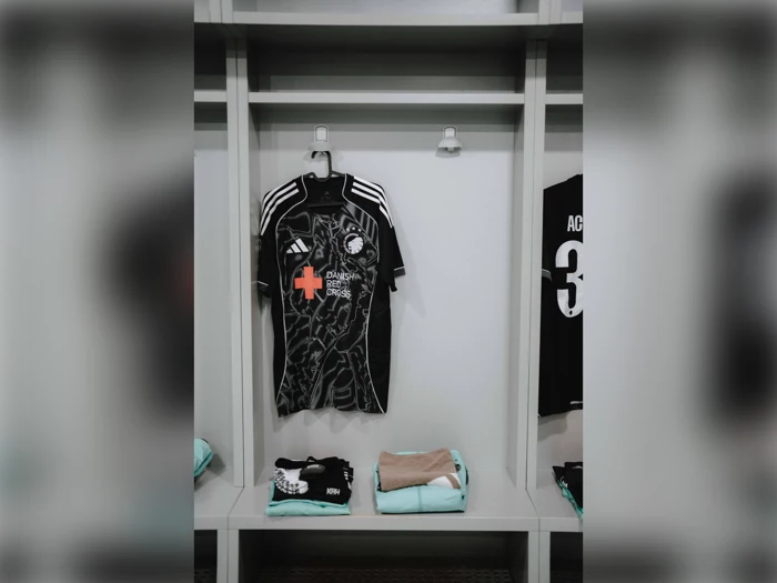 En sort fodboldtrøje med hvide striber og sponsorlogoer hænger i et skab. Sammenfoldet udstyr, herunder lysegrønt tøj, er placeret på hylden nedenunder.