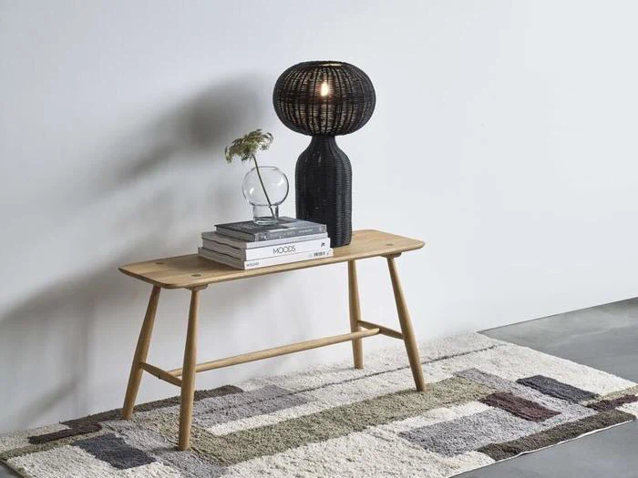 En lille træbænk med stablede bøger, en klar vase med blomster og en sort vævet lampe står på et mønstret tæppe mod en hvid væg.