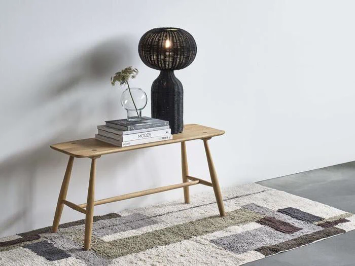 En træbænk med stablede bøger, en rund glasvase med blomster og en sort vævet lampe, alt sammen placeret på et mønstret tæppe mod en hvid væg.