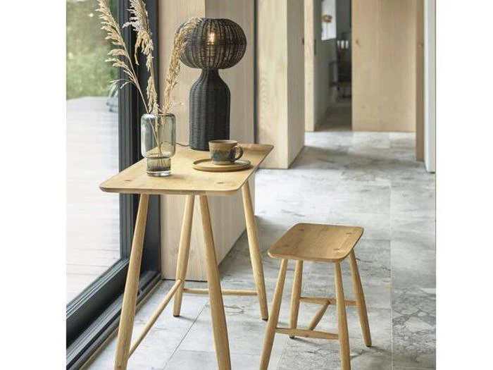 Et lille træbord med en skammel, en vase med tørret græs, en kop og en mørk lampe, placeret ved et glasvindue i et moderne, minimalistisk interiør.