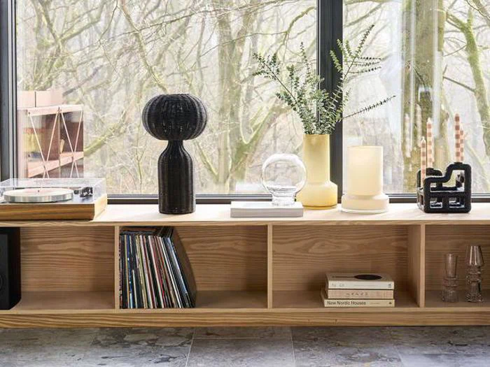 En træhylde med vinylplader, bøger og glas. Ovenpå er der planter, en pladespiller og dekorative genstande ved siden af et stort vindue med udsigt til træer.