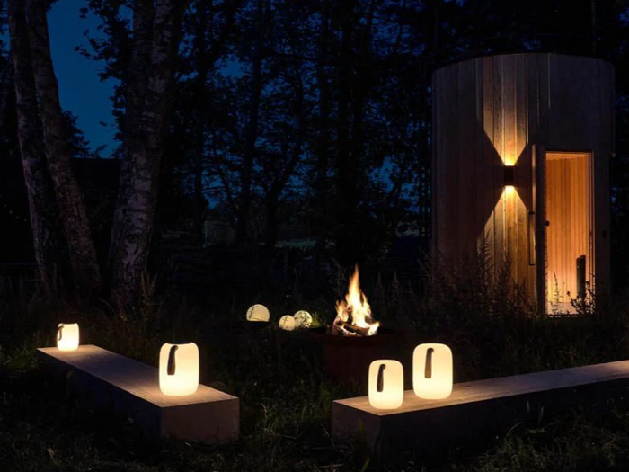 Udendørs scene om natten med lysende lanterner på træbænke, et lille lejrbål og en tændt rund træstruktur i baggrunden blandt træer.