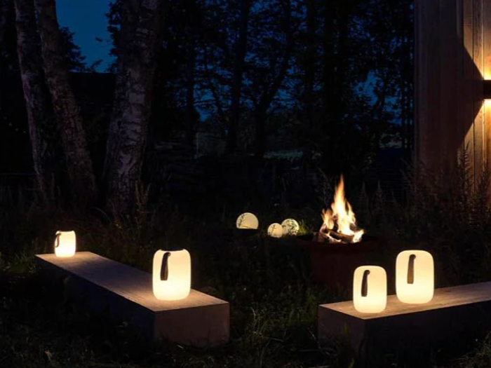 Lysende lanterner på bænke og græs og et bål, der brænder i nærheden, oplyser et udendørs område omgivet af træer om natten.