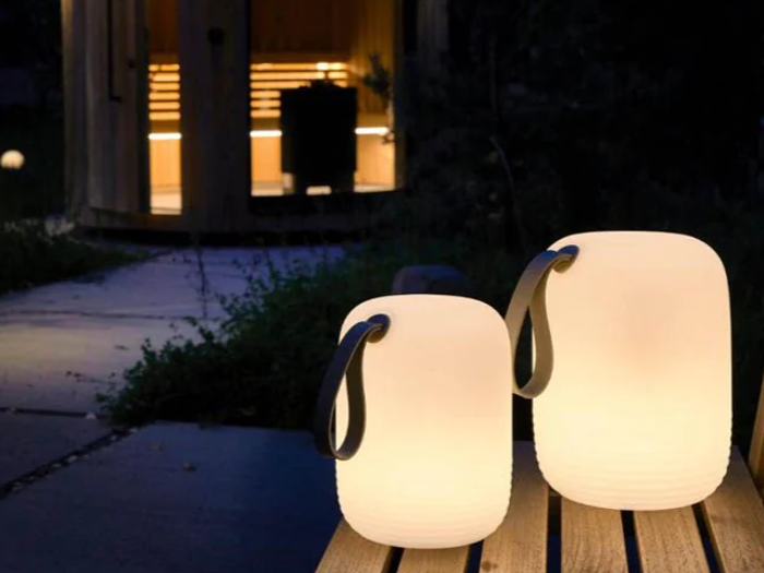 To glødende lanterner med håndtag sidder på en træbænk om natten og oplyser en sti, der fører til en varmt oplyst rund bygning i baggrunden.