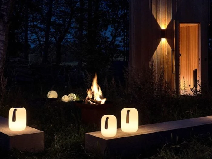 Udendørs scene om natten med et lejrbål, lysende lanterner på bænke og en træbygning oplyst af en væglampe, omgivet af træer.