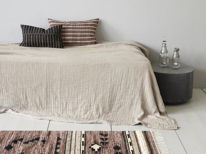 Minimalistisk soveværelse med en pænt redt seng i beige sengetøj, to stribede puder, et rundt gråt sidebord med to glaslanterner og et mønstret tæppe på et hvidt gulv.