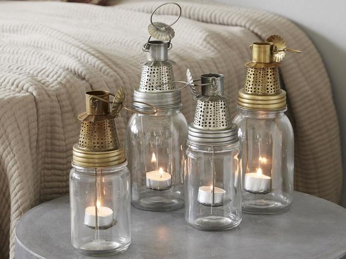 Fire lanterner af glaskrukker med metaltop og tændte fyrfadslys indeni er placeret på et rundt gråt bord; et beige tæppe ses i baggrunden.