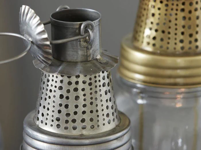 Nærbillede af to vintage lanterner af metal og glas med perforerede toppe; en sølvfarvet i forgrunden og en messingfarvet i baggrunden.