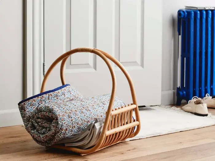 En flettet kurv med hank rummer en sammenrullet blomstret dyne og magasiner, placeret på et trægulv nær en hvid dør og en blå radiator.