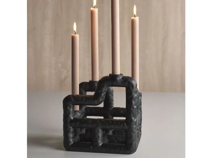 En skulpturel sort lysestage med et geometrisk, industrielt design holder fire tændte beige kertelys på en lysegrå overflade.