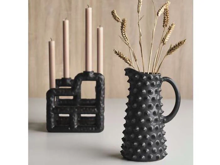 En sort struktureret keramikkande med tørrede hvedestængler ved siden af en matchende lysestage med fire høje beige lys på en lys overflade.