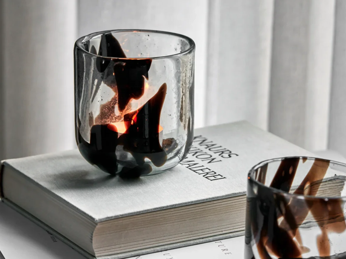 En glaslysestage med sorte hvirvler står på en lukket bog med lysegråt omslag ved siden af et andet lignende glas på en svagt oplyst baggrund.