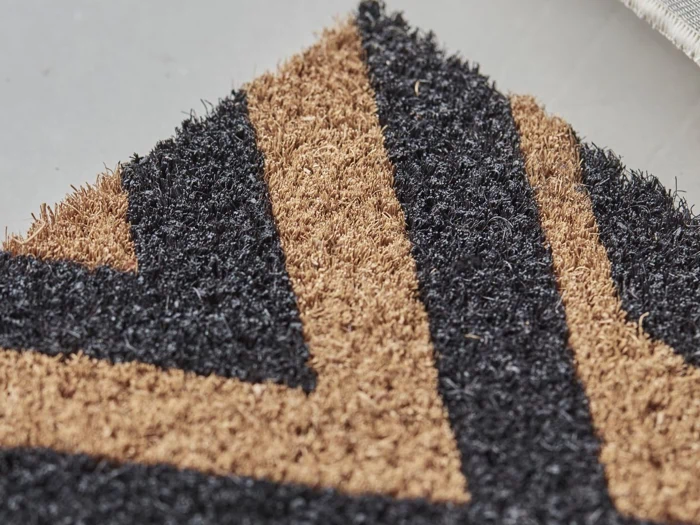 Nærbillede af en dørmåtte med et geometrisk mønster i sorte og brune fibre, der viser et chevron- eller piledesign på en flad overflade.