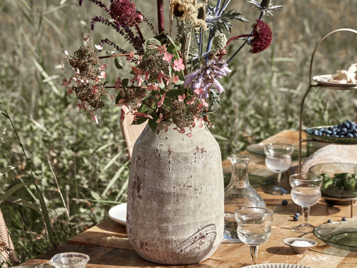 En rustik keramikvase fyldt med forskellige vilde blomster står på et udendørs træbord dækket med glas og tallerkener på en græsmark.