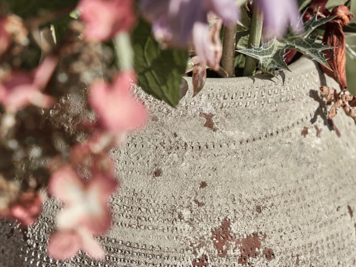 En tekstureret, forvitret keramikkrukke med prikkede mønstre, der holder pink og lilla blomster, vises i nærbillede med en blød, sløret baggrund.