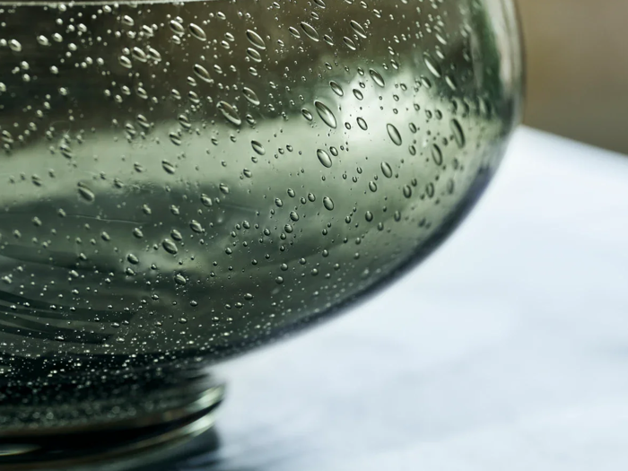 Nærbillede af vanddråber på siden af en gennemsigtig, afrundet glasskål, placeret på en flad overflade med en sløret baggrund.