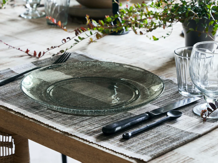 Et spisebord med en klar glastallerken, sort bestik, to klare glas og en dækkeserviet med grønne planter i baggrunden.