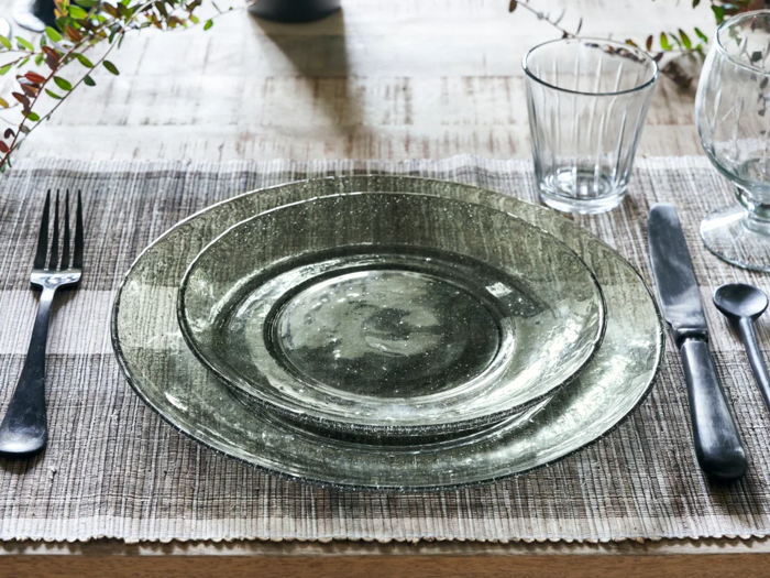 En borddækning med klare glastallerkener, gaffel, kniv, ske og et glas på en vævet dækkeserviet, dekoreret med grønne blade.