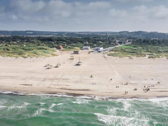 En bred sandstrand med blide bølger, et par både og mennesker spredt langs kysten og en lille kystby med grønne områder i baggrunden under en delvis overskyet himmel.