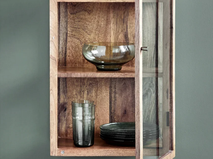Et vægskab i træ med en glasdør, der viser en glasskål, et glasglas og en stak mørke tallerkener på to hylder.