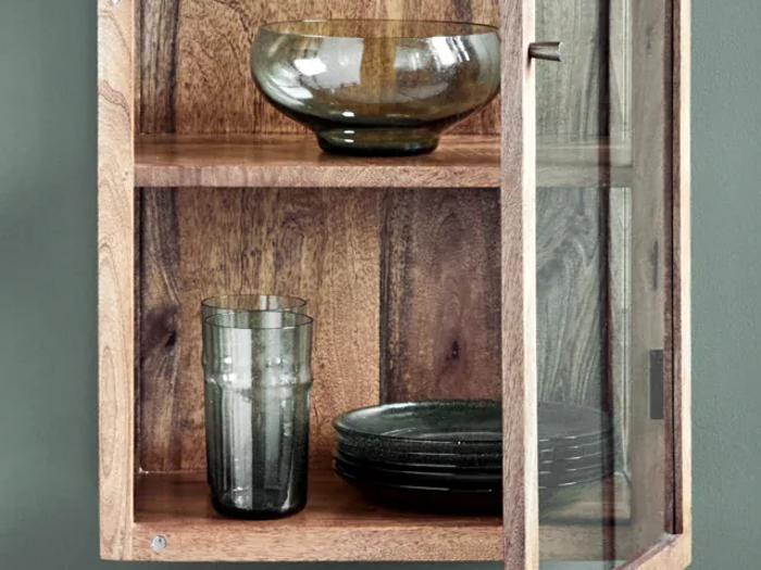 I et træskab med glasdør står en glasskål på den øverste hylde og flere mørke glastallerkener og et glas på den nederste hylde.