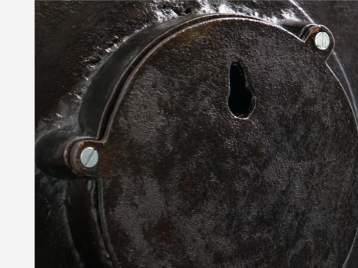Nærbillede af en rusten, rund metalplade med to skruer og en nøglehulsformet åbning nær den øverste midte.