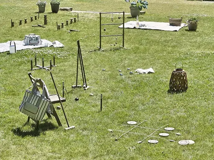 Udendørs græsplæne med forskellige havespil, bl.a. tic-tac-toe, stigekast, og spredt udendørs indretning og picnic-artikler på græsset.