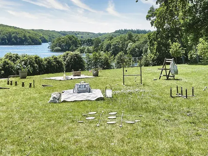 Et græsareal ved en sø med picnictæpper, kurve, tallerkener og plænespil opstillet under træer med frodigt grønt og vand i baggrunden.