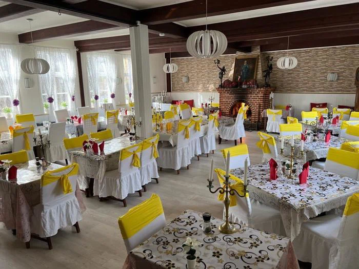 En lys spisestue med mange borde dækket af duge, gule stolesløjfer, røde servietter og dekorative kandelabre, med store vinduer og en muret pejs.
