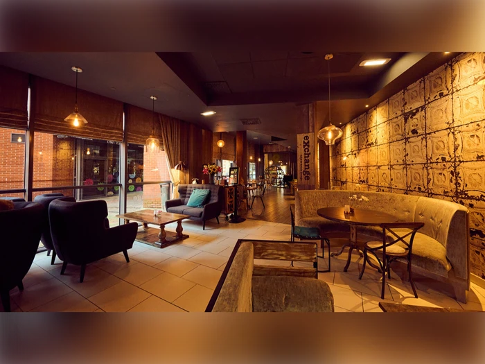 En hyggelig, svagt oplyst café med bløde stole, sofaer, runde og firkantede borde, varm belysning og store vinduer, der viser den udvendige murstensvæg.