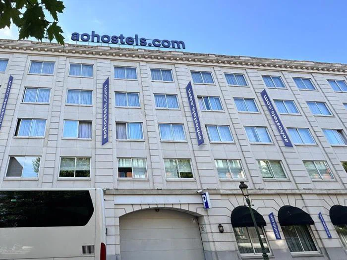 En hvid bygning i flere etager med blå "aohostels.com"-skilte på taget og mellem vinduerne, og en hvid bus parkeret foran.